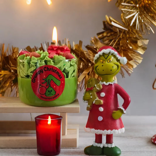 Grumpy Christmas geurkaars van ToHotToCandle® groene sojawas kaars met geur van den en slingers, naast kerstfiguur en rode decoratie